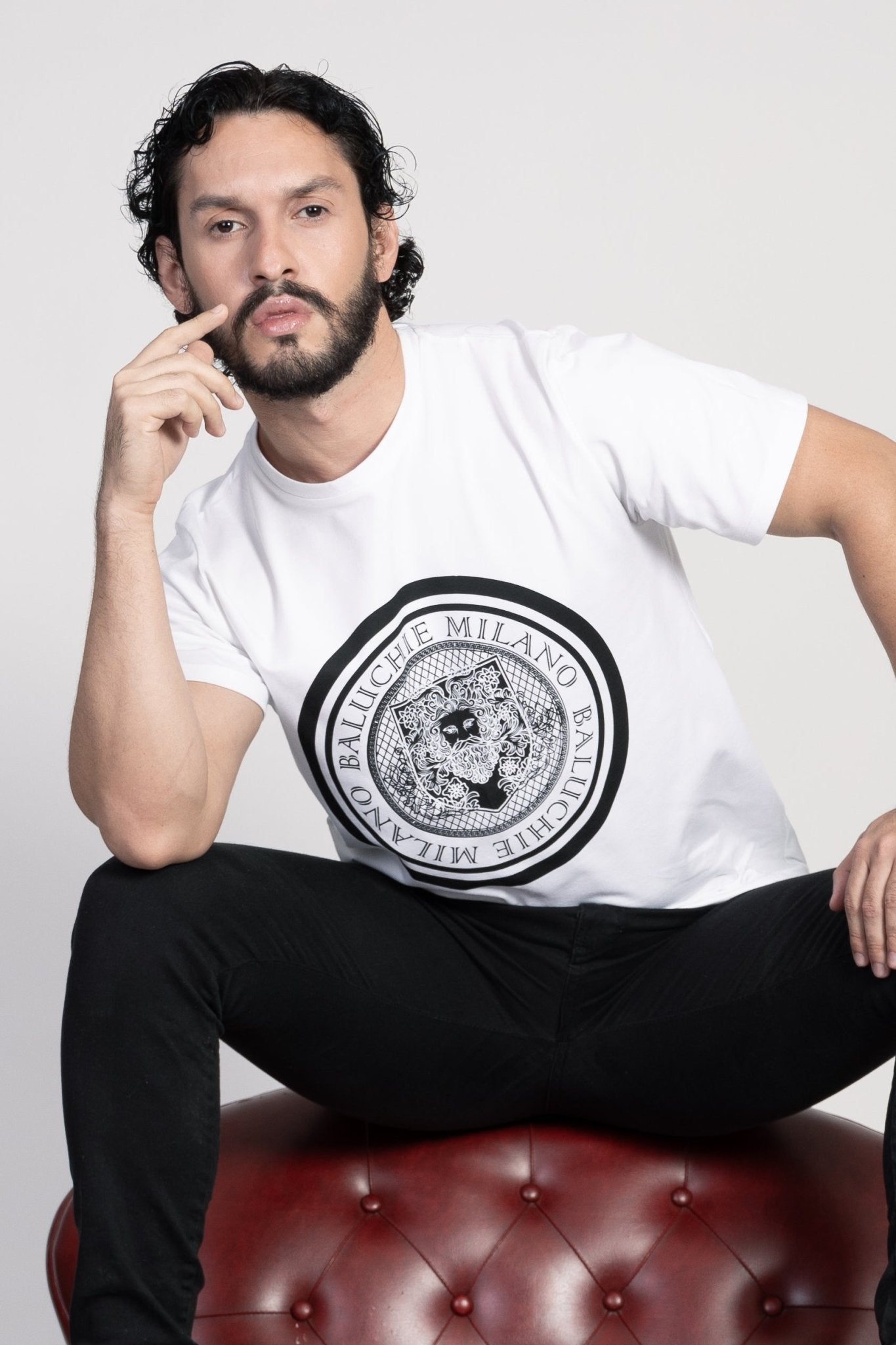 Man looking forward at the camera, Baluchoe Milano coin logo t-shirt. Also, sitting on the couch. 