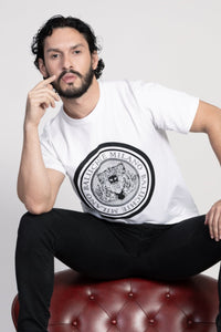 Man looking forward at the camera, Baluchoe Milano coin logo t-shirt. Also, sitting on the couch. 